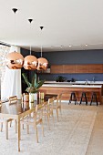 Pendant lamps with copper-coloured lampshades above dining set with long designer table and chairs made from pale wood; kitchen with free-standing counter in background