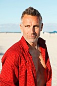 Mann mittleren Alters mit rotem, aufgeknöpftem Hemd am Strand