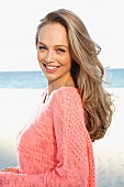 A young blonde woman by the sea wearing a pink summer jumper