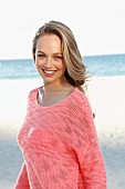 A young blonde woman by the sea wearing a pink summer jumper