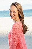 A young blonde woman by the sea wearing a pink summer jumper