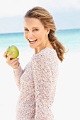 A blonde woman on a beach wearing a long, knitted jumper holding an apple