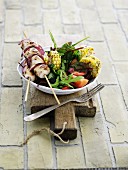 A chicken skewer with red onions, grilled corn on the cob and a salad