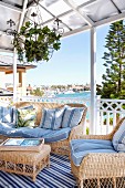 Helle Rattan Sitzmöbel mit hellblauen Polstern auf überdachter Terrasse, im Hintergrund Meeresküste