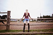 Blonde langhaarige Frau in Karohemd und knappen Shorts steht an Holzzaun, Rückansicht