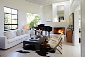 Chairs with sheepskin blankets and white sofa around coffee table; grand piano next to open fireplace in background in living room