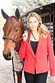 A young blonde woman wearing a red jacket next to a horse