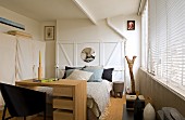 Schreibtisch vor Bett an weisser Holzwand in rustikal modernem Schlafzimmer, seitlich geschlossene Jalousie am Fenster