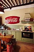 Wooden kitchen in pale vintage green with retro advertising signs, Coca-Cola barrels and old workbench used as counter