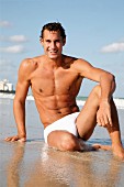 Young, athletic man in swimming trunks sitting on sandy beach