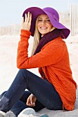 A young blonde woman sitting in the sand wearing an orange cardigan and a purple hat