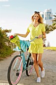 A young blonde woman pushing a bicycle wearing a yellow summer dress