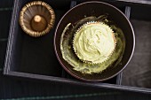 A cupcake with matcha tea frosting in a bowl with matcha tea powder next to a tea whisk