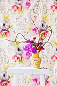 Anemonen, Ranunkeln und Orchidee in gelb geblümter Vase vor floral gemusterter Tapete