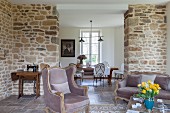 Sofa and armchair in modernised, rustic interior with stone wall and view of dining area through floor-to-ceiling open doorway