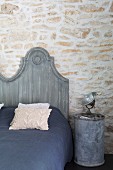 Grau gestrichenes Bettkopfteil an rustikaler Natursteinwand, Blechtonne als Nachttisch mit Strahler
