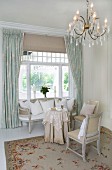 Kronleuchter über eleganter Sitzecke mit zierlichen, weissen Antikmöbel vor traditionellem, skandinavischem Fenster mit Vorhang