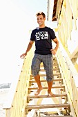 Junger Mann mit T-Shirt und karierter Hose auf eine Holztreppe am Strand