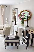 Armchair and footstool with black and white striped upholstery next to bouquet in front of antique, gilt-framed mirror on vintage console table