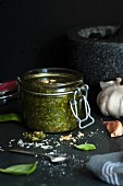 A jar of homemade basil pesto with ingredients and a mortar