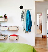Teilweise sichtbares Bett mit grüner Tagesdecke, gegenüber an Wand montierte Holzboxen und Hakenleiste mit Tüchern, neben offener Tür