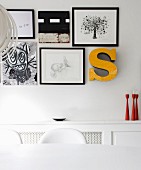 Black and white pictures, drawings and yellow ornamental letter above red candlesticks on white shelf