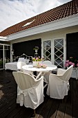 Chairs with white loose covers at round table on wooden terrace outside house with black and white façade