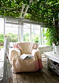 Comfortable armchair on wooden floor in front of white-painted wooden lattices with roller blinds on veranda with climber-covered pergola