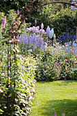 Blick durch Metall Rankbogen auf blaue Veronika und blühenden Stauden, dazwischen Gartendeko