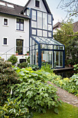 Büsche in verschiedenen Grüntönen im Garten, im Hintergrund Wohnhaus mit Wintergarten, blaue Stahl- Glaskonstruktion