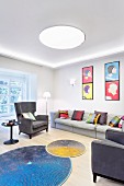 Brightly coloured scatter cushions on grey sofa and armchairs below pop-art-style pictures and round colourful rugs in front of fireplace