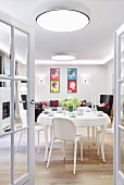 View through open glass double doors of white dining area with plastic chairs and pop-art pictures above seating area in background