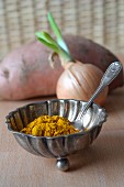 Turmeric powder in a silver dish with an onion and a sweet potato in the background