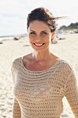 A brunette woman on a beach wearing a beige, openwork jumper