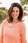 A brunette woman on a beach wearing a salmon-coloured top and an openwork jumper