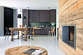 Fireplace in rustic wooden wall; dining area with antique table and modern, charcoal grey kitchen counter in background