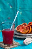 A glass of blood orange juice with a straw with a plate of juiced blood oranges in the background