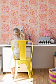 Girl sitting on yellow-painted metal chair at desk against wall with retro floral wallpaper
