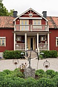 Ornaments and rusty metal candle holders in flowerbed in front of Falu-red country house with porch and balcony