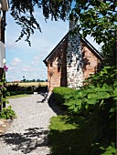 Sonnenbeschienener Kiesweg im Garten, im Hintergrund schlichtes Backsteinhaus mit Aussenkamin