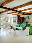 Delicate tray tables in front of sofa with white loose cover and scatter cushions in various shades of green in open-plan interior with dining area in background