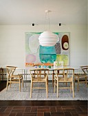 Dining set with pale wooden classic chairs on rug below white designer pendant lamp in front of modern artwork on wall