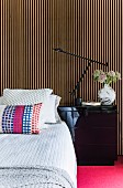 Kissen auf Bett mit Tagesdecke, seitlich schwarzer Nachttisch vor Wand mit Holzlamellenverkleidung