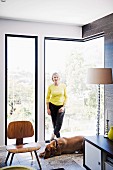 Woman wearing yellow top and dog in front of frameless corner window, replica of classic chairs and postmodern standard lamp with plexiglas base