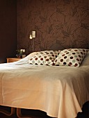 Polka-dot pillows and pale blanket on double bed against brown wallpaper with floral pattern
