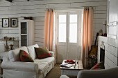 Sofa with scatter cushions next to French windows with curtains in corner of living room with white wood-clad walls