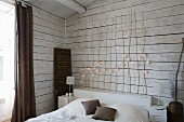 Simple bed with wooden headboard made of branches decorated with fairy lights against white, wood-clad wall