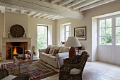 Wicker armchairs, sofa and coffee table in front of fireplace in renovated country house with whitewashed wood-beamed ceiling