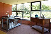 Kunsthandwerkliche Anrichte und Edelstahlspüle mit Milchkannen und Kochtopf in spärlich möbliertem Raum mit Fensterfront