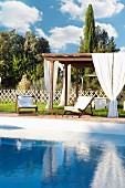 Holzpergola mit weißen Vorhängen und Holzliegestühle mit weißen Polstern im sommerlichen Garten am Pool